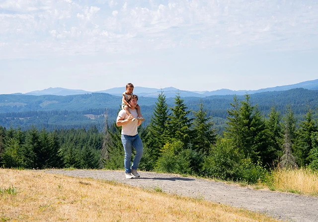A father carrying his son on a hiking trail with trees and mountains in the background